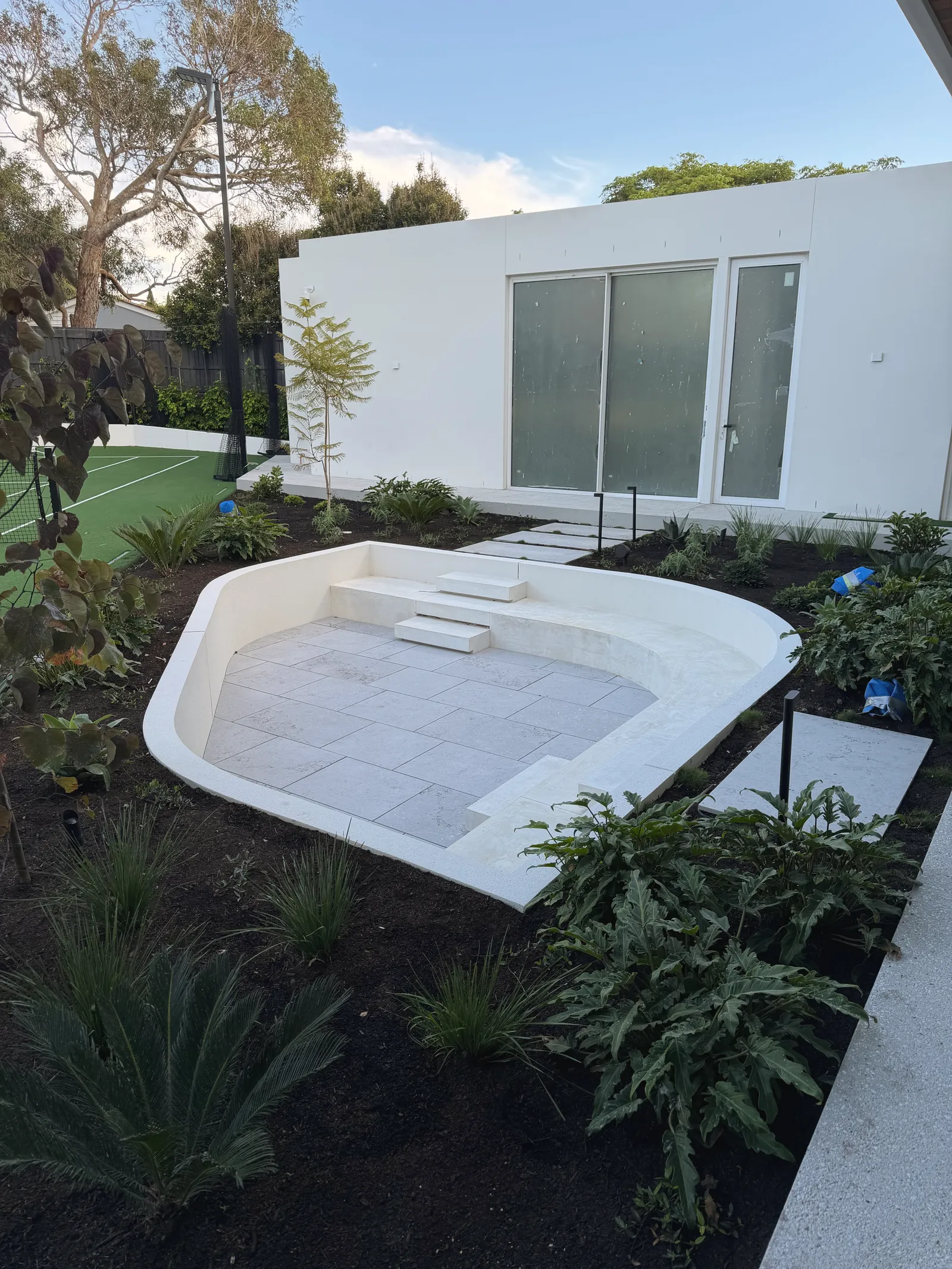 Curved rendered sunken seating area with pale stone paving, set into a planted garden beside a white rendered house