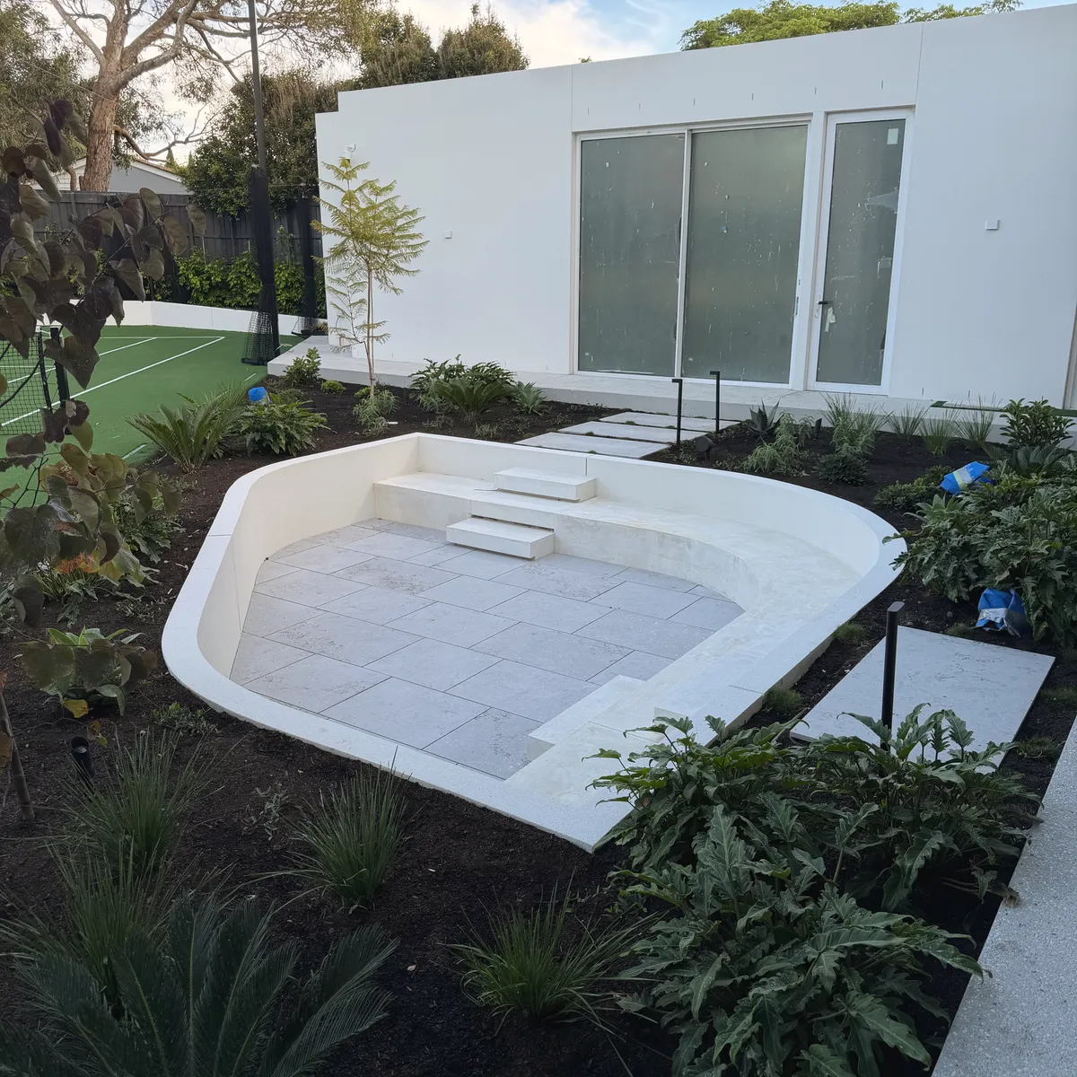 Curved rendered sunken seating area with pale stone paving, set into a planted garden beside a white rendered house
