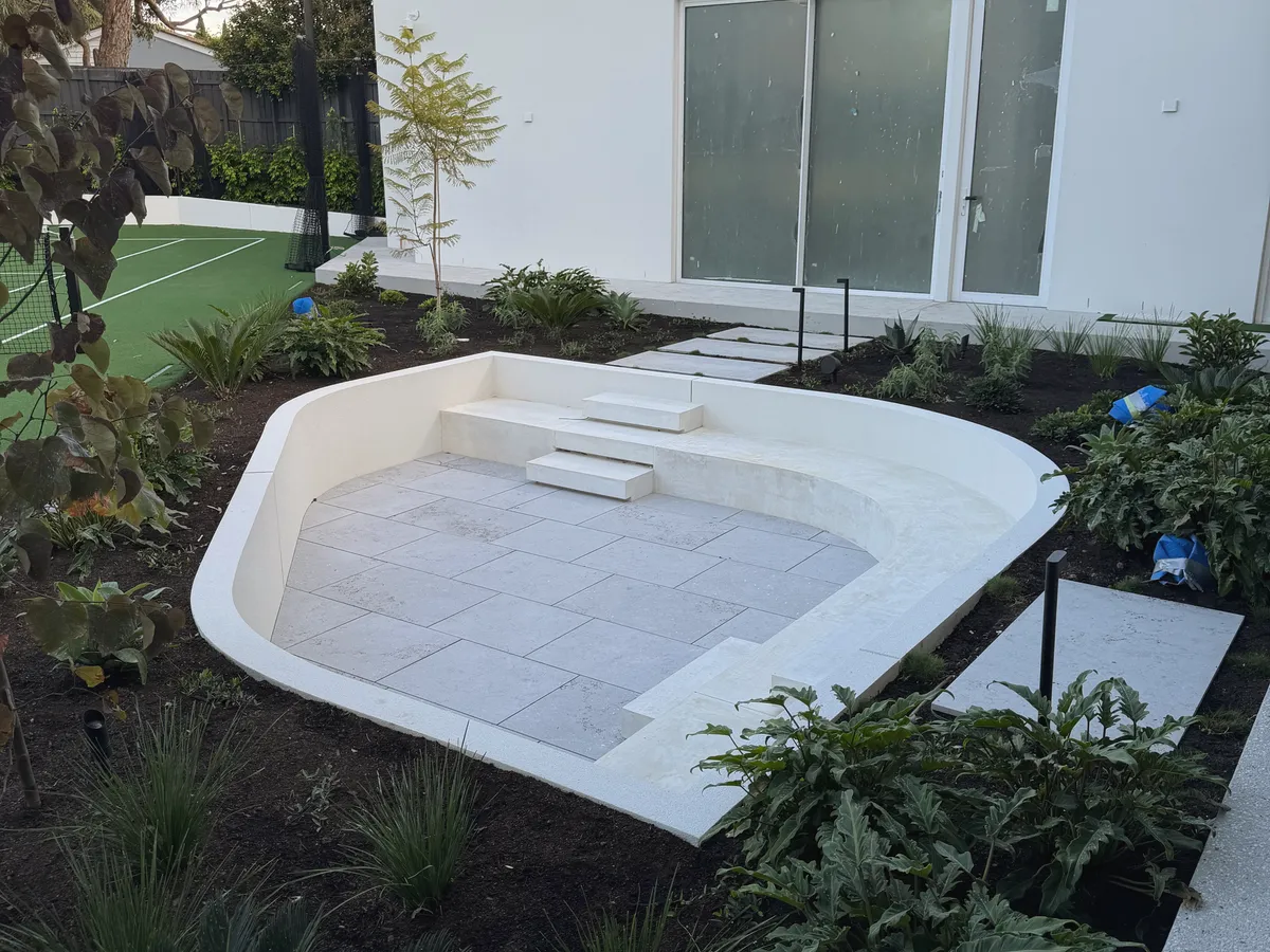 Curved rendered sunken seating area with pale stone paving, set into a planted garden beside a white rendered house