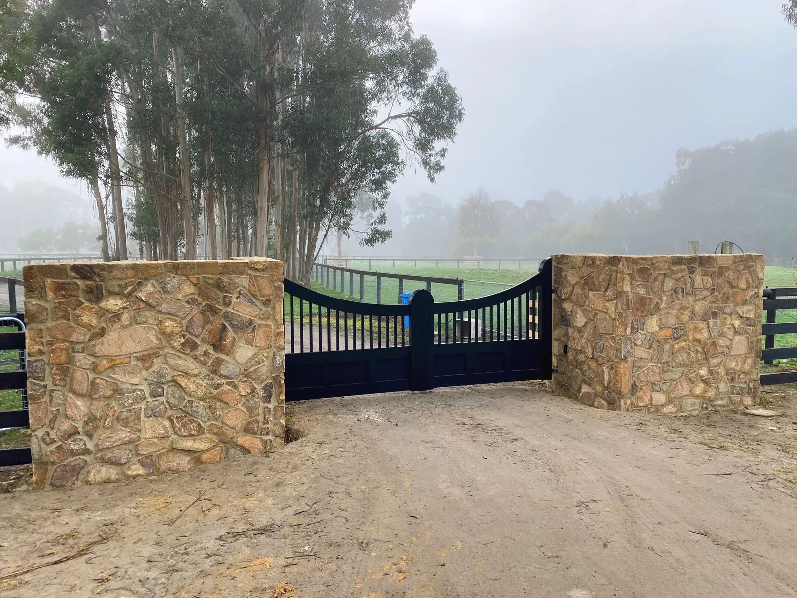 Stone-clad gate pillars and a black gate at a rural entrance on a foggy morning, post-and-rail fencing either side