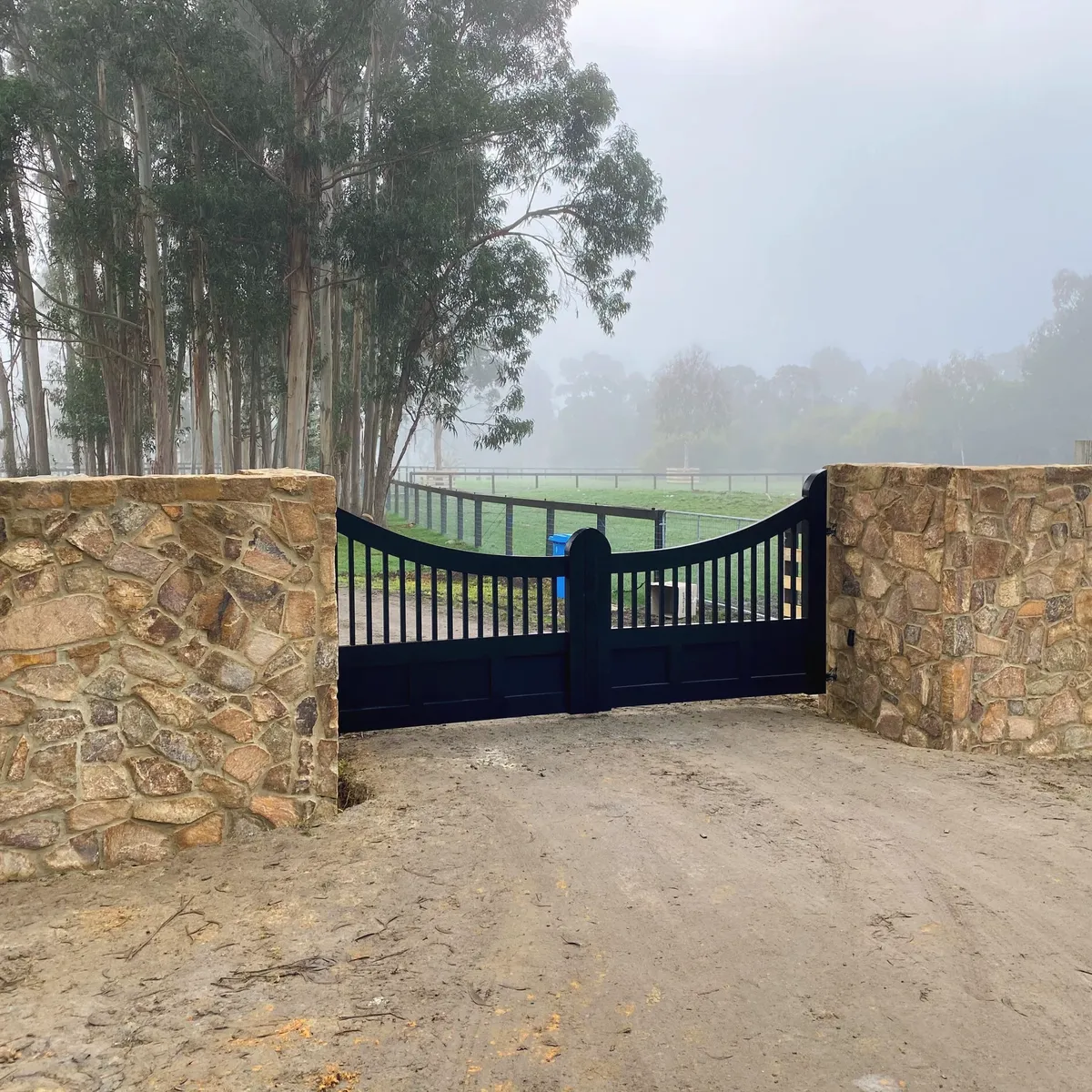 Stone-clad gate pillars and a black gate at a rural entrance on a foggy morning, post-and-rail fencing either side