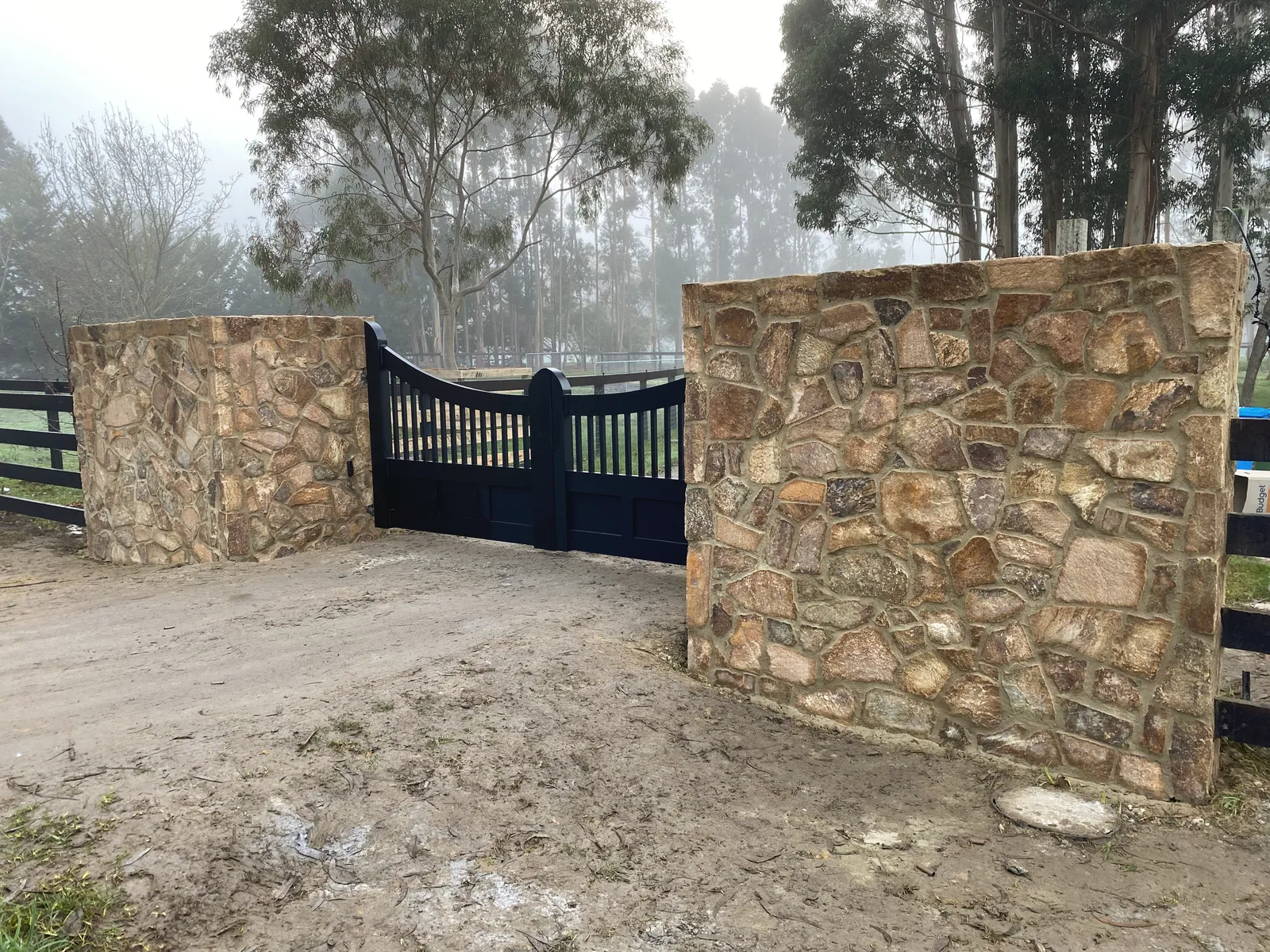 Two stone-clad entrance pillars either side of a black timber farm gate on a rural driveway