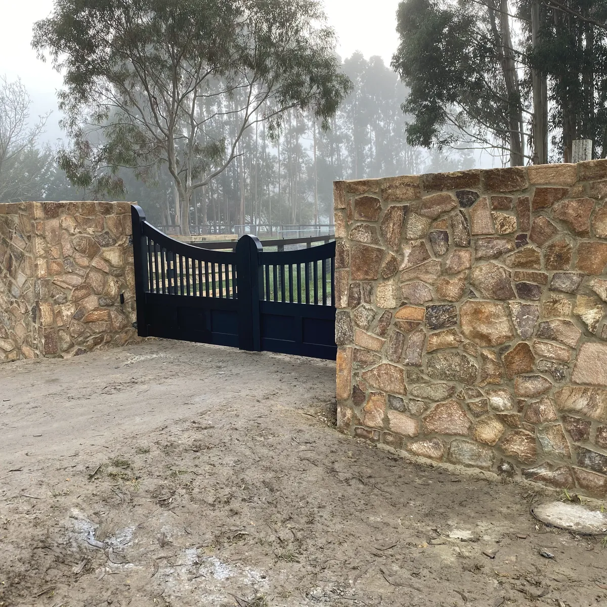 Two stone-clad entrance pillars either side of a black timber farm gate on a rural driveway