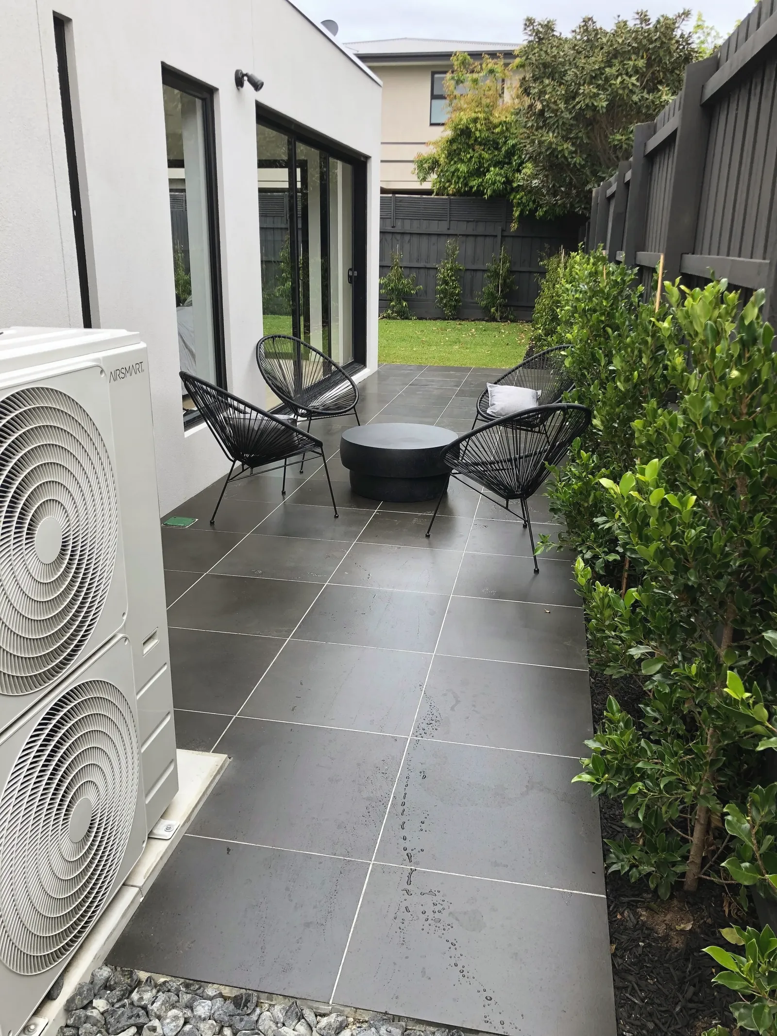 Charcoal large-format paving in a narrow side courtyard, with a fire pit, wire chairs and a new hedge