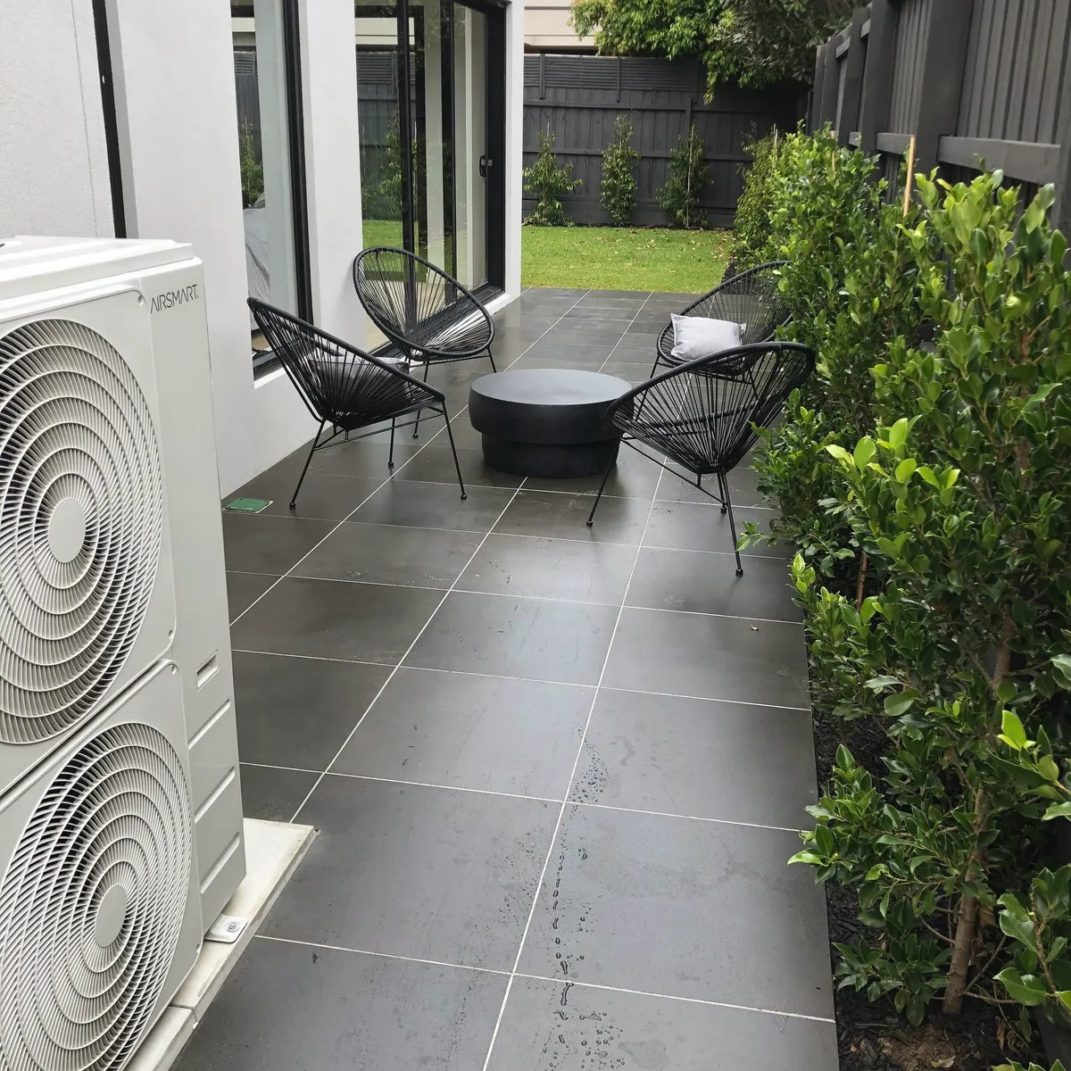 Charcoal large-format paving in a narrow side courtyard, with a fire pit, wire chairs and a new hedge