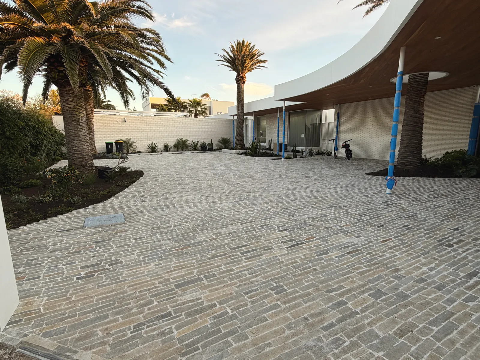 Wide cobblestone-paved courtyard and driveway laid in a running bond, mature palms and a curved white building behind