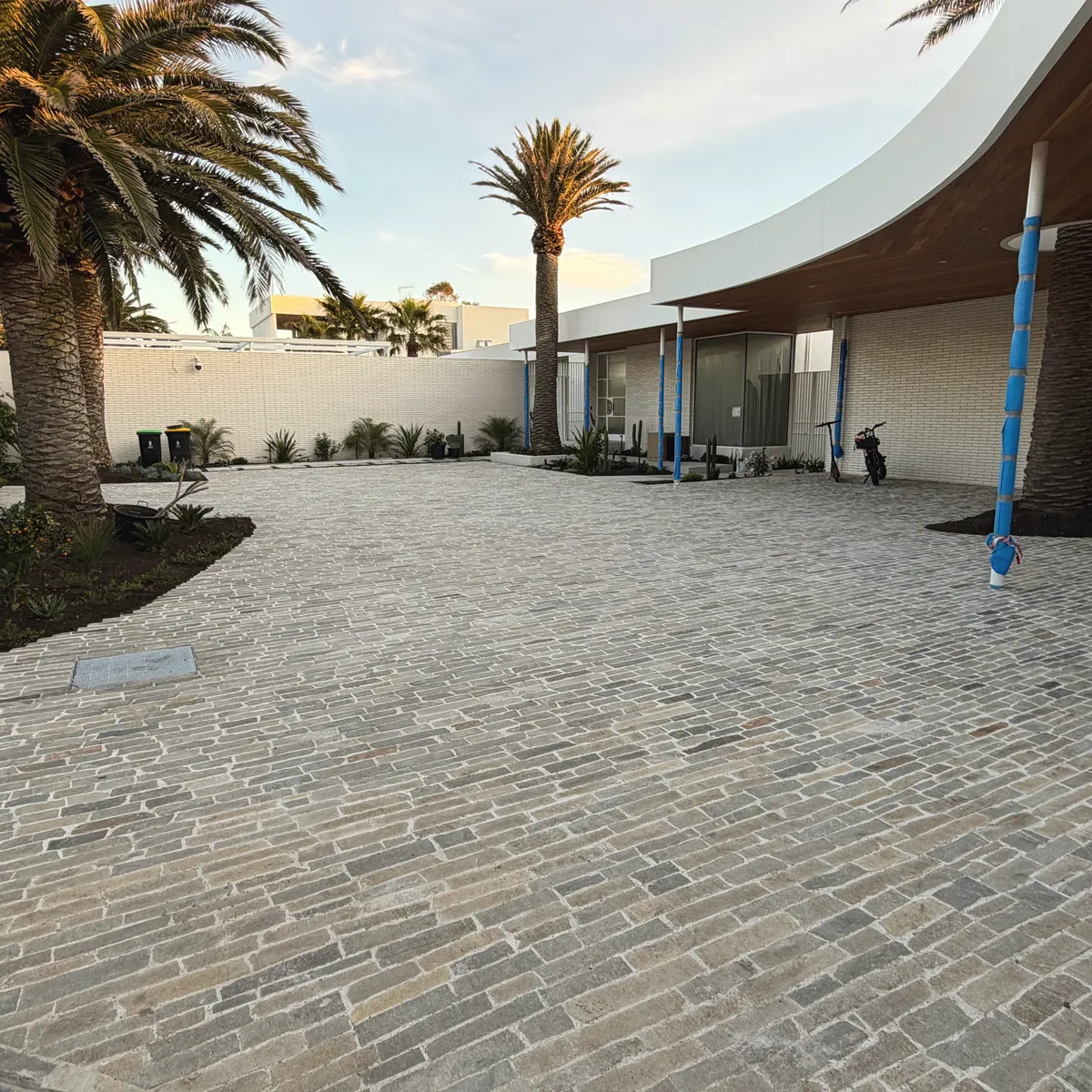 Wide cobblestone-paved courtyard and driveway laid in a running bond, mature palms and a curved white building behind
