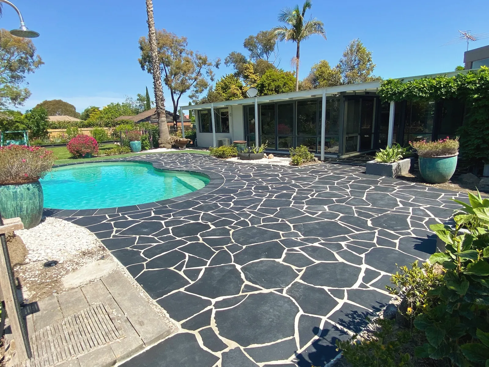 Charcoal crazy paving laid around a kidney-shaped pool, with potted plants and a mid-century house behind