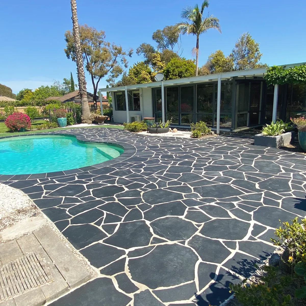 Charcoal crazy paving laid around a kidney-shaped pool, with potted plants and a mid-century house behind