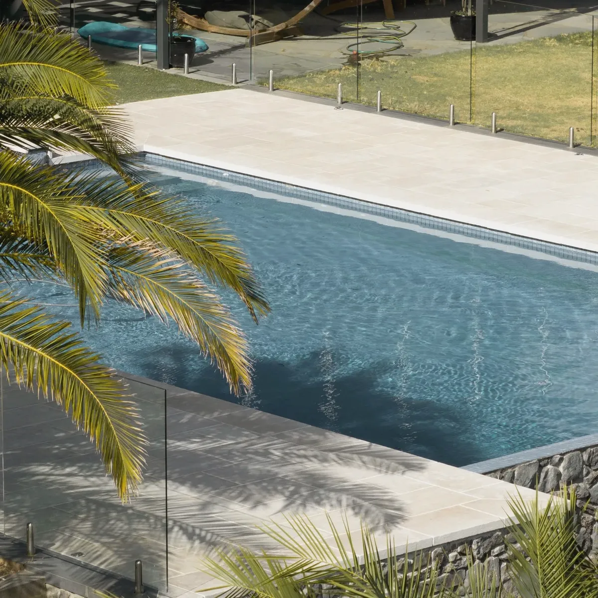 Pool with pale stone coping and a frameless glass fence, a dry-stone wall along the lower edge