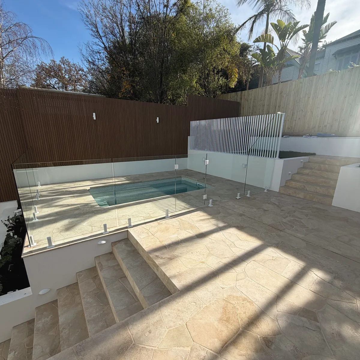 Swimming pool with a frameless glass fence and a flagstone travertine surround, steps rising to a rendered upper terrace