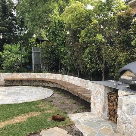 Curved stone seating wall with a timber bench, pizza oven and flagstone patio under festoon lights