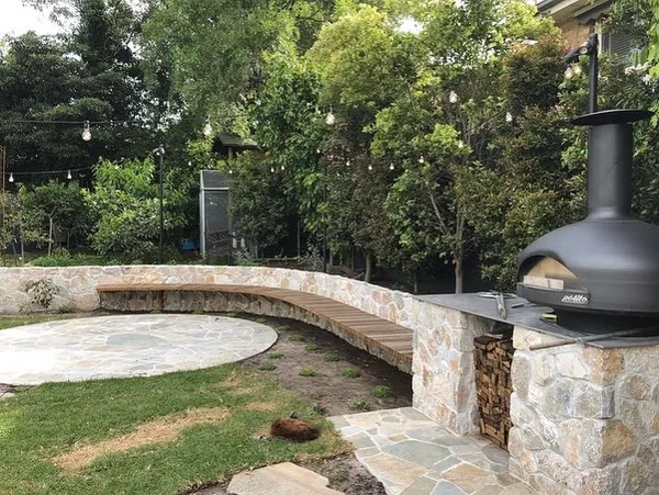 Curved stone seating wall with a timber bench, pizza oven and flagstone patio under festoon lights